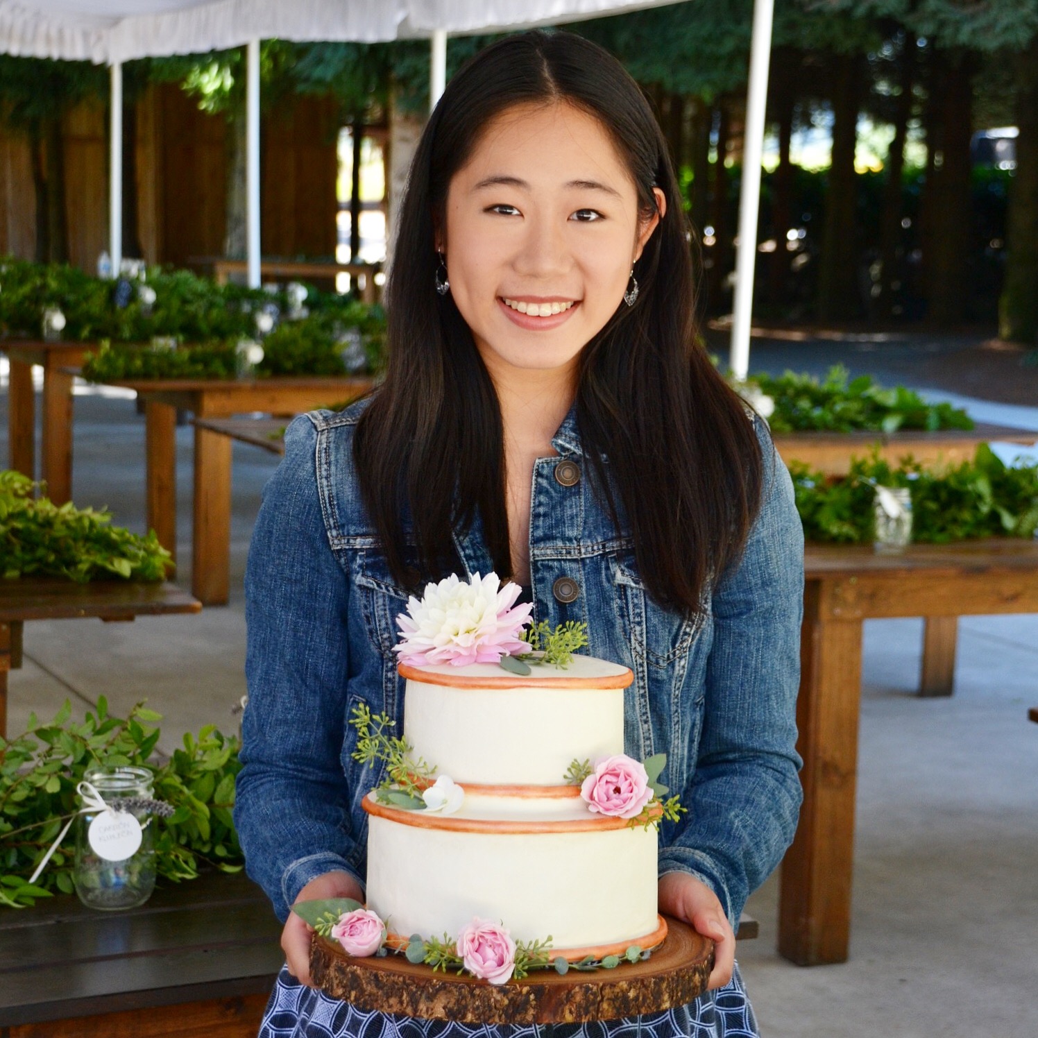 Lucy, a Chinese American woman, holds a two-tiered wedding cake with pastel pink flowers and rose gold detailing.