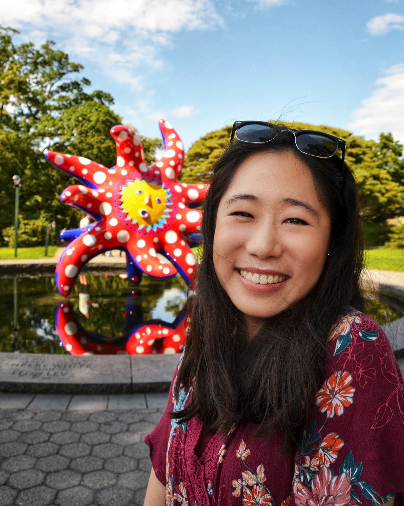 Headshot of Lucy, a Chinese American woman, wearing a floral maroon romper and standing in front of a whimsical red and yellow Yayoi Kusama statue.