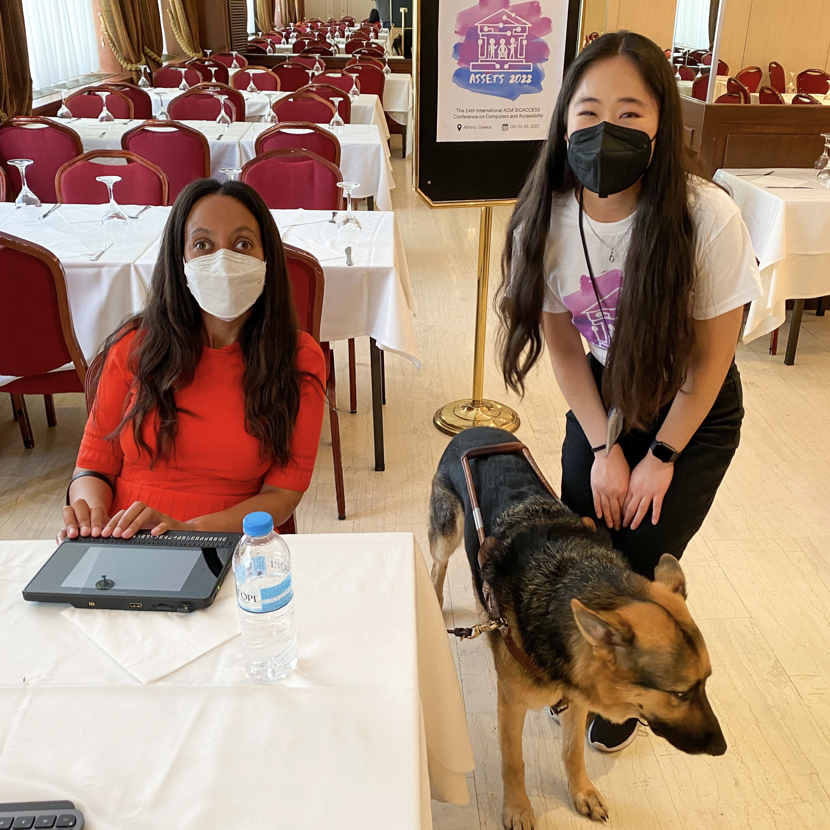 Haben Girma wears a red dress and sits at a table with her Braille display. Lucy, wearing an ASSETS shirt, stands next to her. Both are wearing masks. Mylo, Haben's Seeing Eye dog, stands between them.