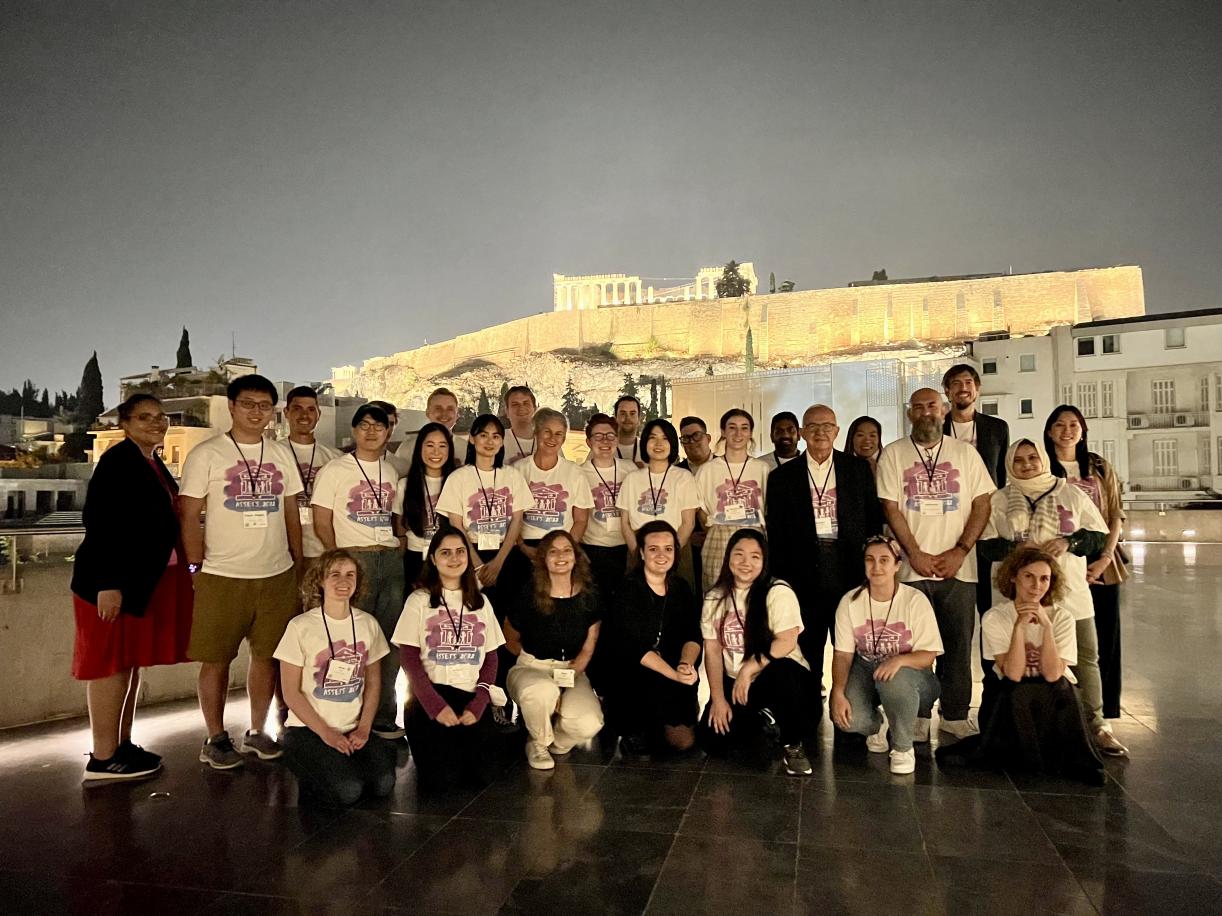 The ASSETS Organizing Committee and Student Volunteers wearing the 2022 shirt stand outside at night with the Acropolis and Parthenon in the background.