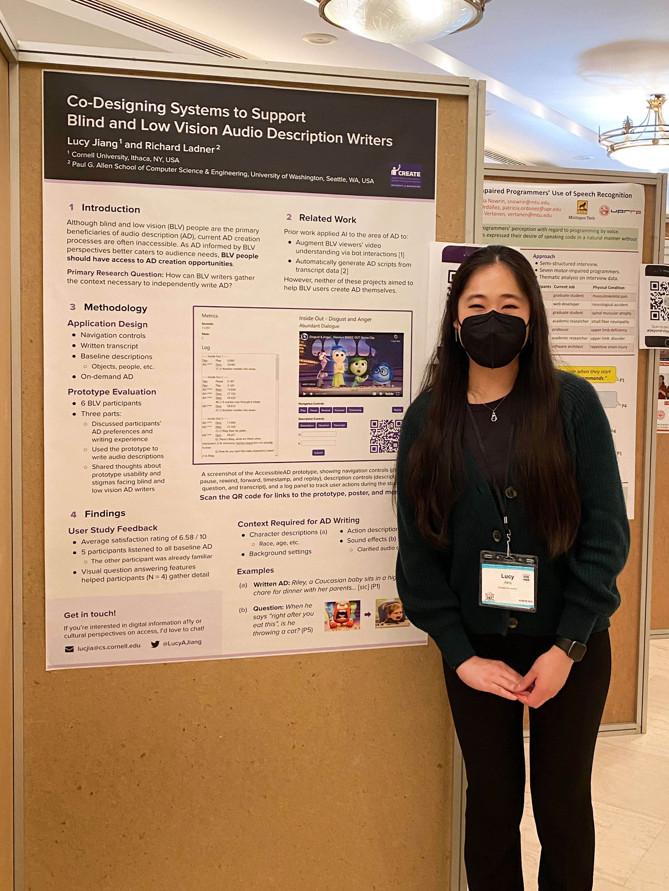 Lucy is smiling and wearing a dark green sweater, black dress pants, a silver penguin necklace, and a black face mask. She stands to the right of her ASSETS poster, which is taped on the wall.