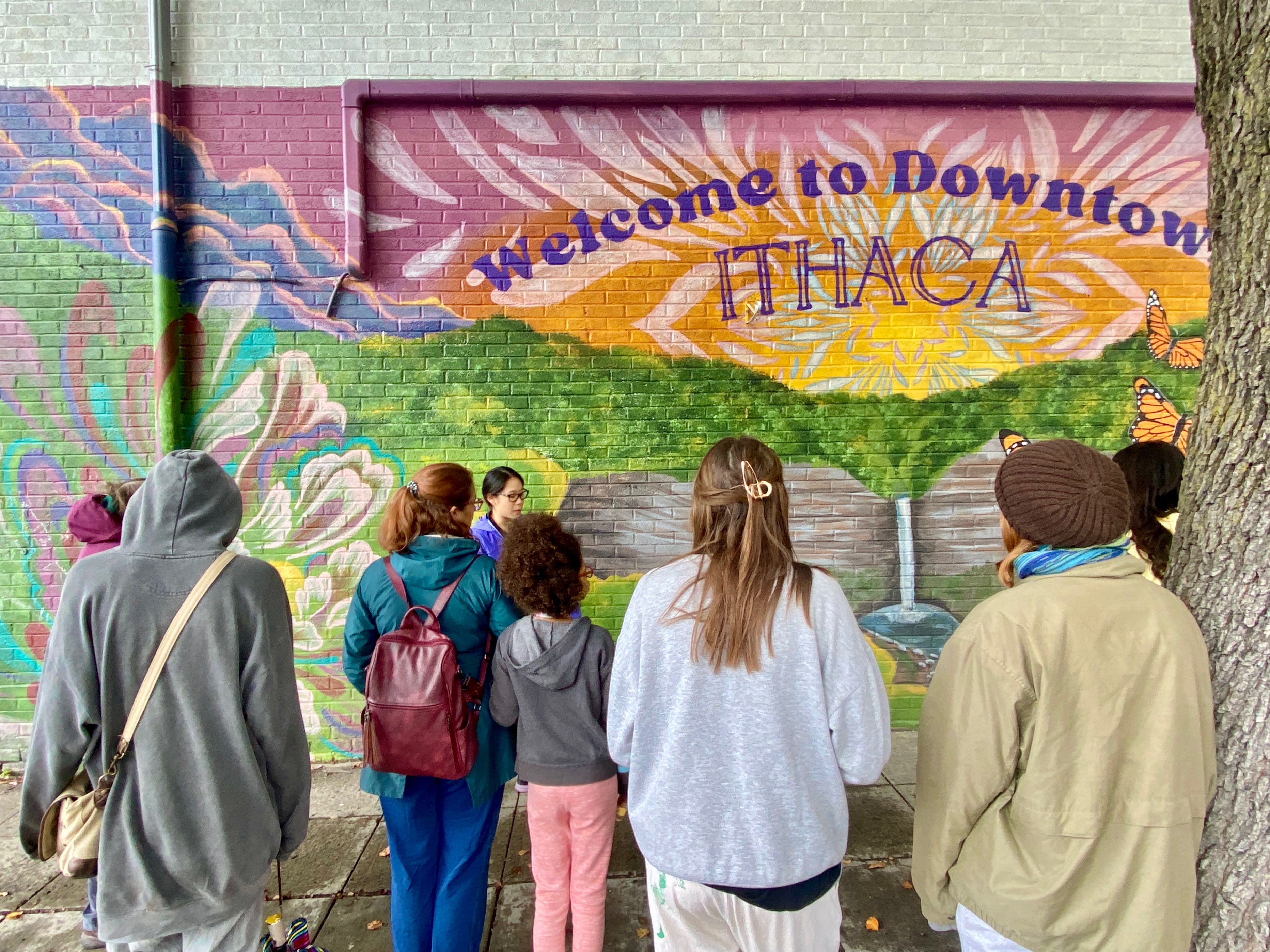 Seven mural tour attendees stand facing a mural with their backs to the camera. The mural is called 'Welcome to Downtown Ithaca' and was painted by Kaitlyn Cronin. The mural shows a large, vibrant sunset over Taughannock Falls (a tall, thin waterfall) with abstract flowers on the left and larger-than-life monarch butterflies flying upwards on the right. The mural says 'Welcome to Downtown' in an arc and 'Ithaca' below it. Lucy stands in front of the mural, reading a description.