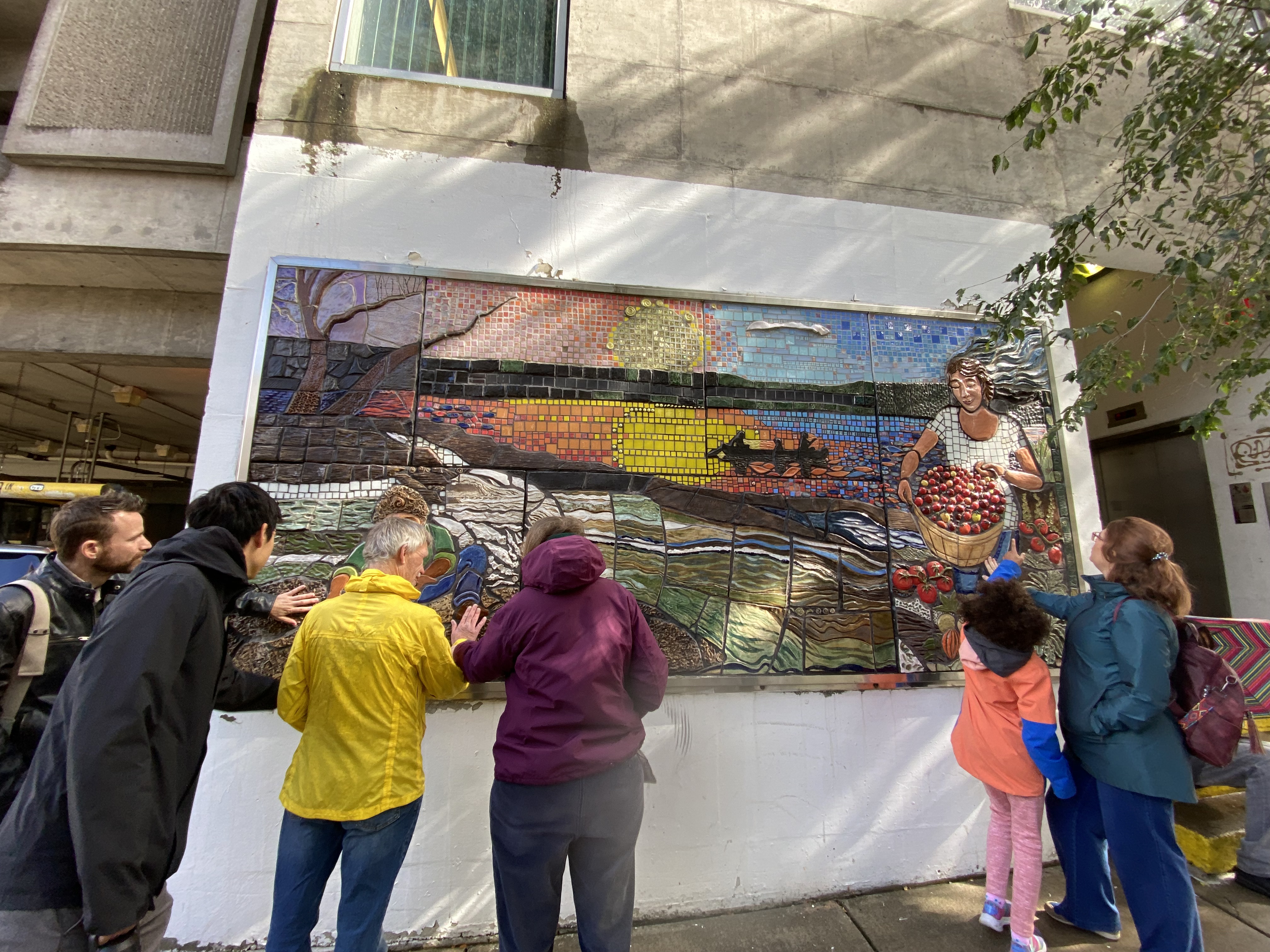 Six mural tour attendees wearing colorful rain jackets reach out to feel the textured tiles near the bottom of a large mosaic. Their backs are to the camera. The mosaic is called 'Feels Like Home - Spirit of Ithaca' and was created by Annemarie Zwack. The mosiac shows a sunset scene with a child planting seeds on the left, a woman carrying a bushel of tomatoes on the right, and three canoers in the background. The tiles on the mosaic are different sizes, shapes, and textures; towards the bottom there are large ceramic tiles with a ripple texture, whereas the sky and water are composed of small, smooth tiles. The tomatoes in the bushel are also three-dimensional.