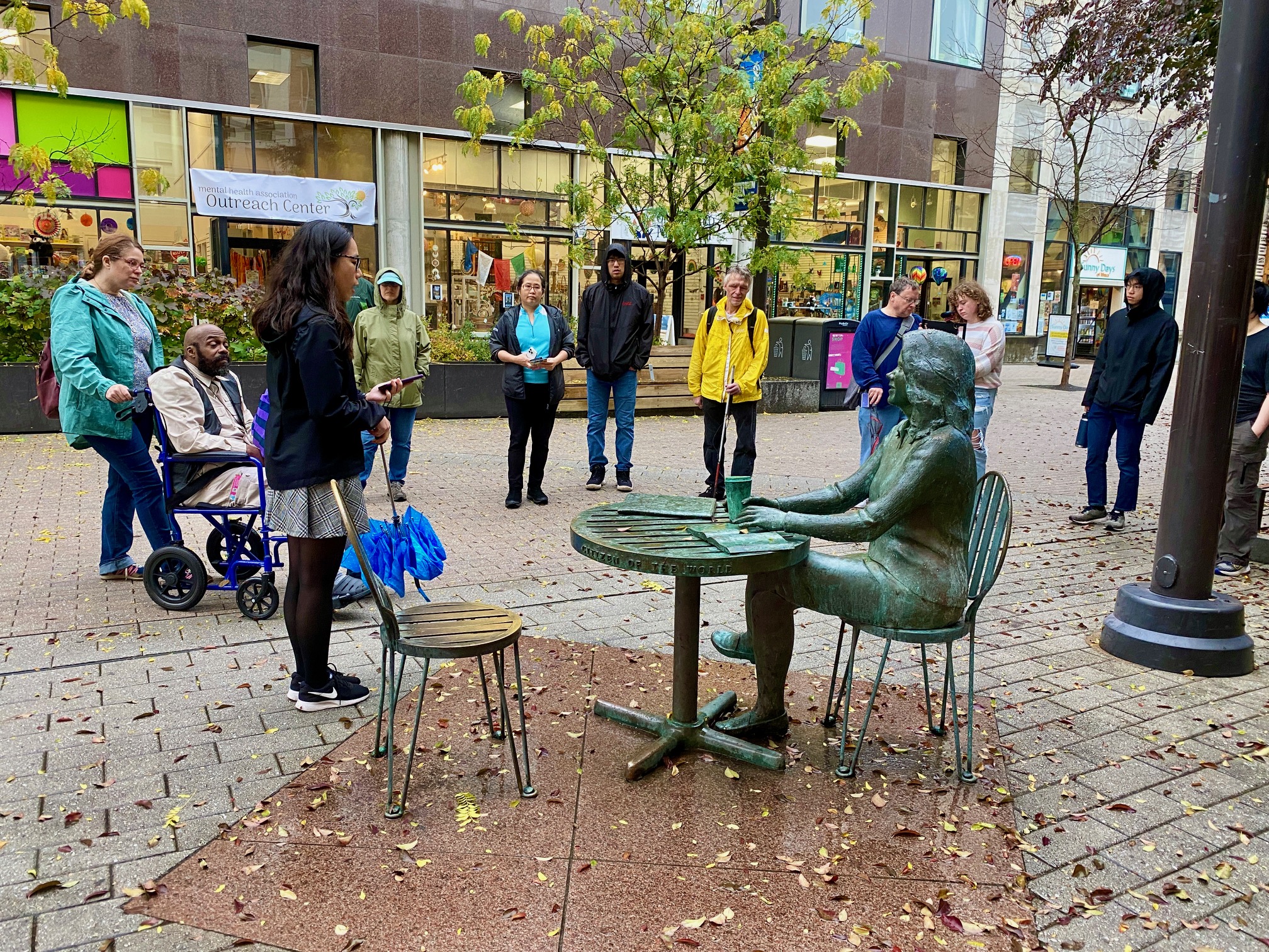 Ten mural tour attendees are gathered in a circle around the 'Child of Ithaca' sculpture. The group is facing the camera and the back of the sculpture is shown. The sculpture is a life-size bronze sculpture of a young woman with shoulder-length hair sitting at a table, with a coffee cup in her right hand and an open book before her. There is another chair opposite from the woman. The sculpture has a green patina over its entire surface; only the seat across from the woman retains its bronze color. One attendee is a wheelchair user and one holds a white cane. Lucy stands by the empty chair in the sculpture, holding an umbrella and reading out a description from her phone.